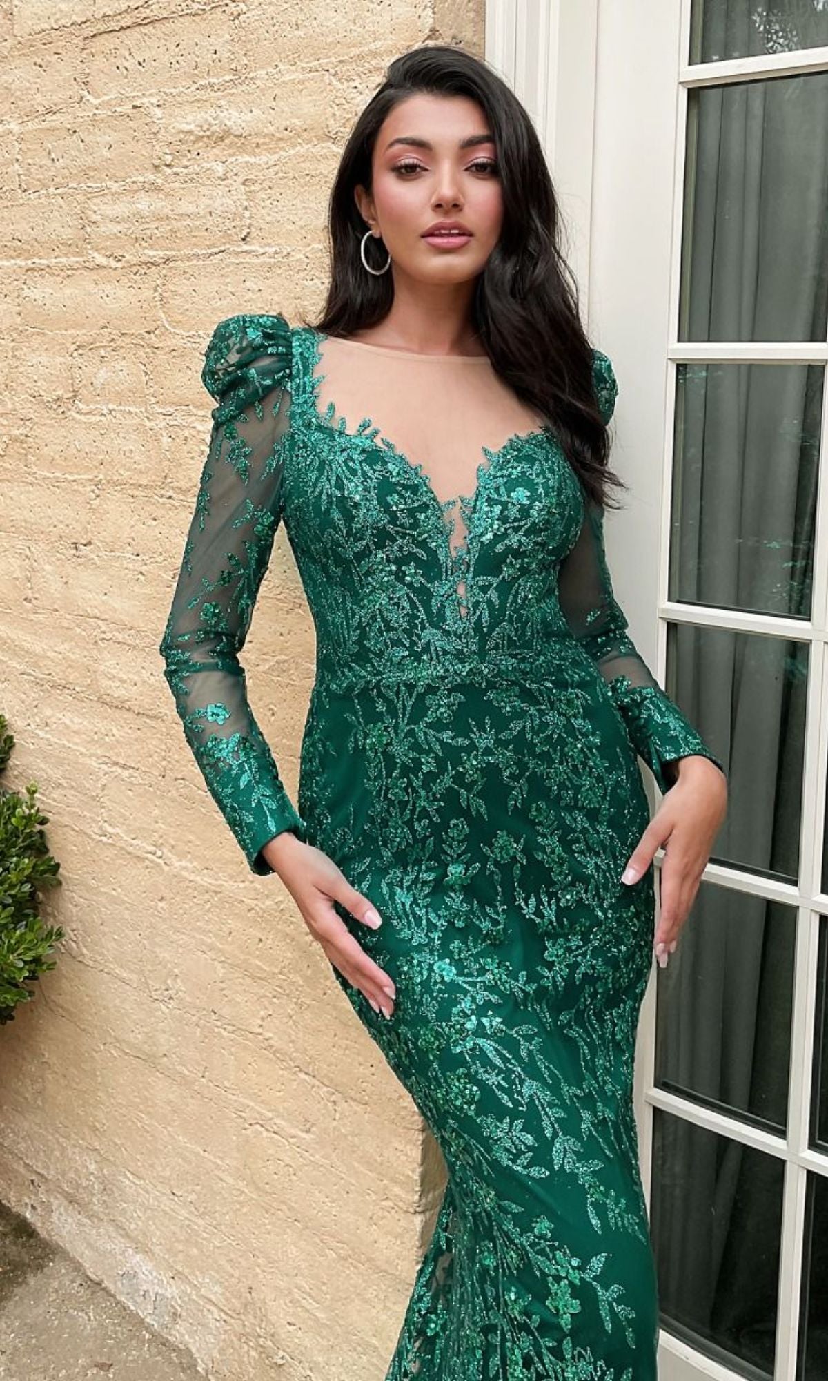 Woman wearing the Long Sleeve Long Glitter-Lace Prom Dress CD989 in green, featuring a fitted silhouette and sheer details, standing before a beige wall and glass-paneled door.